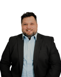 A professional headshot of a man with short dark curly hair and a beard, wearing a light blue shirt and a dark suit jacket. He is smiling and facing the camera against a plain white background.