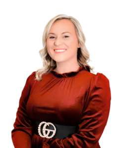 A professional headshot of a woman with blonde hair, wearing a red dress with a black belt featuring a pearl-encrusted buckle. She is smiling and facing the camera against a plain white background.