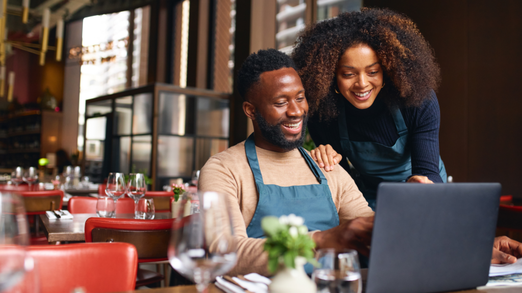 A smiling couple, wearing aprons, looking at a laptop in a cozy restaurant setting with red chairs and set tables in the background.