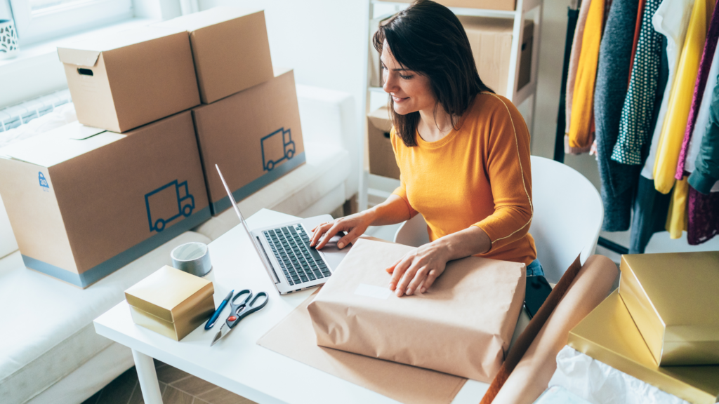 A woman wearing a yellow shirt is working on a laptop at a table surrounded by packing materials, boxes, and clothing.