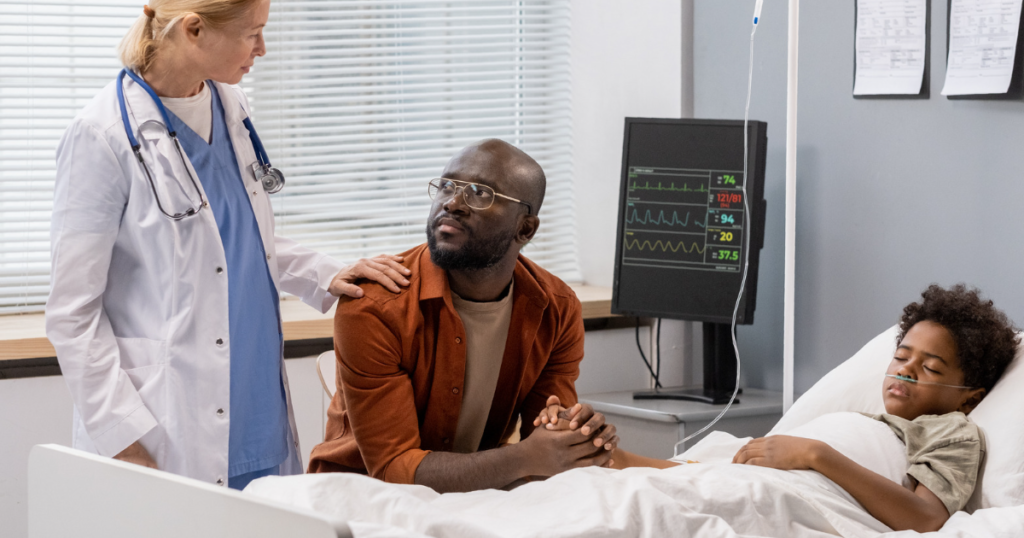 A concerned father sits at the bedside of his child, who is resting in a hospital bed with a nasal cannula. A doctor, wearing a white coat and stethoscope, stands beside them, gently placing a hand on the father's shoulder, offering reassurance.